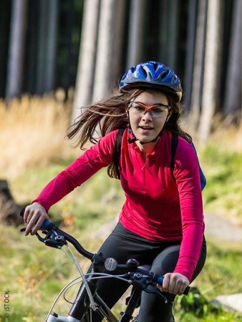 Una donna che indossa una camicia rossa a maniche lunghe, un casco e degli occhiali sta andando in bicicletta in una zona erbosa e boschiva in una giornata di sole.