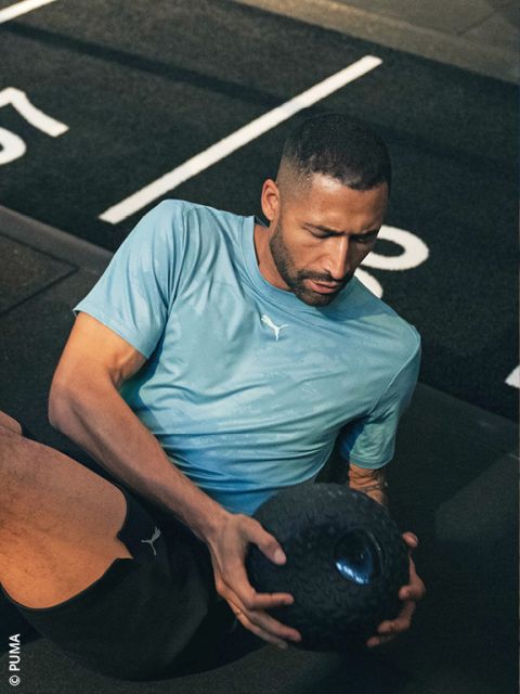 Un uomo con una maglietta Puma azzurra si allena sul pavimento di una palestra, tenendo con entrambe le mani una palla medica nera e strutturata.