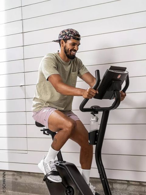 Un uomo che indossa un cappello a fiori, una t-shirt beige e pantaloncini rosa sorride mentre pedala su una cyclette Schwinn.