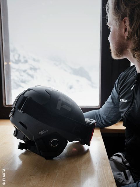 Una persona con capelli chiari e barba è seduta a un tavolo di legno vicino alla finestra e guarda fuori verso le montagne innevate. Un casco da sci nero è appoggiato sul tavolo davanti a loro.