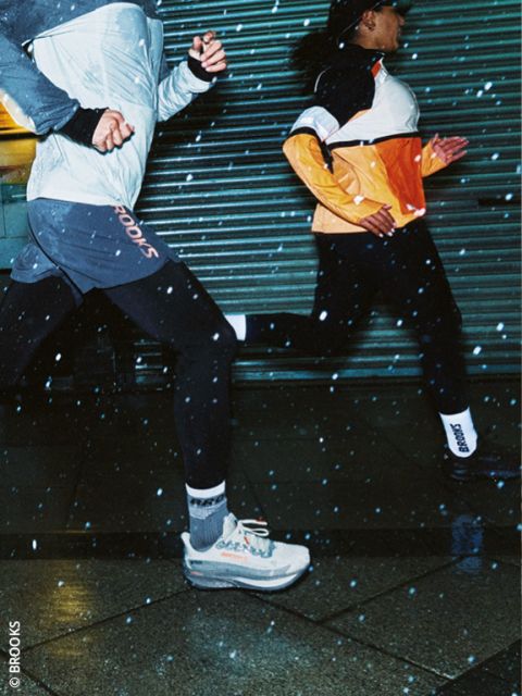 Due persone che corrono di notte in abbigliamento sportivo, con gocce di pioggia visibili nell'aria e marciapiede bagnato sotto di loro. Entrambi i corridori indossano giacche e scarpe da corsa e corrono velocemente davanti a un cancello di metallo.