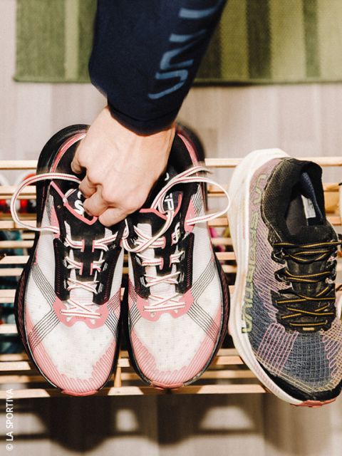 Una persona tiene in mano un paio di scarpe da corsa bianco-rosa per i lacci sopra una scarpiera di legno, accanto a una singola scarpa da corsa nera e grigia.