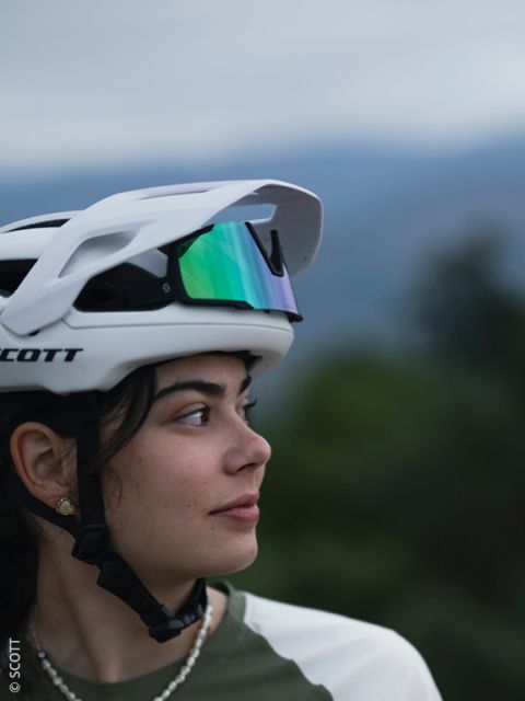 Una giovane donna con un casco da bici bianco e occhiali da sole riflettenti sulla testa guarda di lato. Lo sfondo è sfocato con verde naturale e un cielo morbido e nuvoloso.
