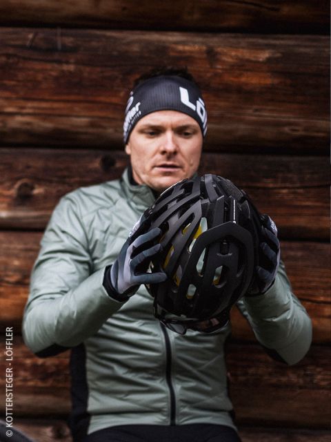 Un uomo con giacca e guanti verdi esamina un casco da bicicletta nero ed è seduto davanti a una parete di legno. Indossa una fascia nera con scritte bianche.