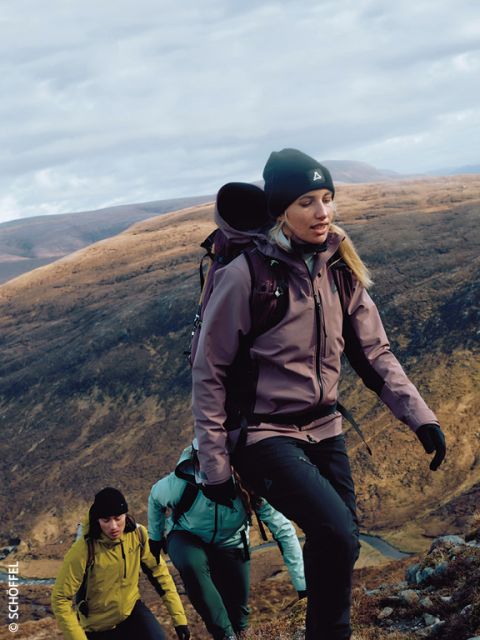 Tre escursionisti con attrezzatura da esterno e zaini scalano un pendio roccioso con dolci colline marroni sullo sfondo sotto un cielo nuvoloso. L'escursionista in testa indossa una giacca viola e un berretto.