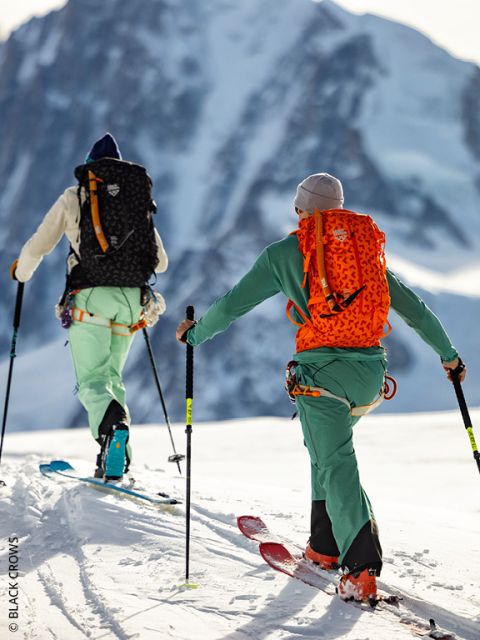 Due persone in colorati abiti da sci e zaini salgono su una montagna innevata, aiutandosi con i bastoncini. Sullo sfondo si vede una grande e aspra cima montuosa.