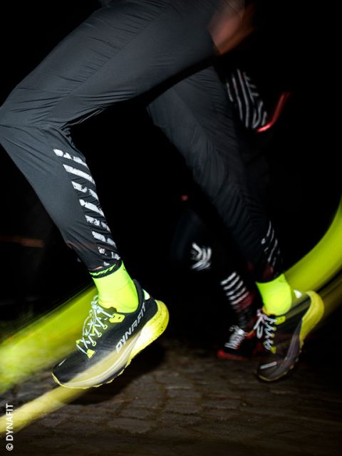 Un primo piano di due persone che corrono di notte. L'attenzione principale è sulle loro gambe, che indossano pantaloni sportivi neri, calze giallo neon e scarpe da corsa DYNAFIT gialle e nere, con la sfocatura del movimento che enfatizza la velocità.