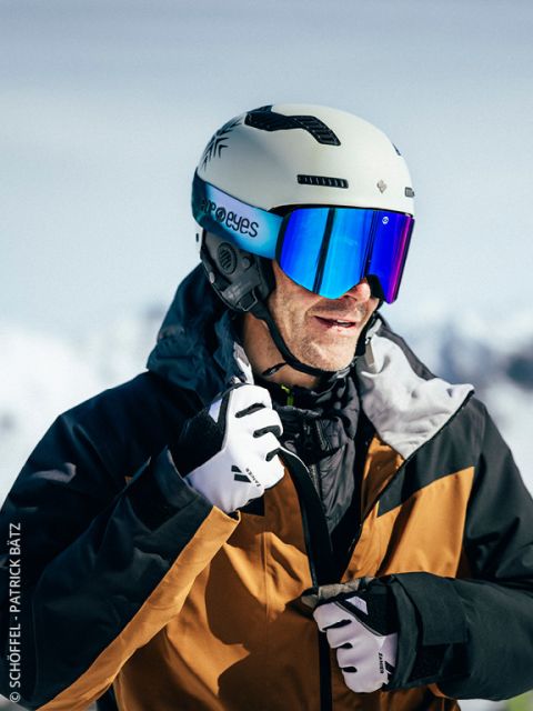 Uno sciatore che indossa un casco bianco, occhiali riflettenti, una giacca e guanti marrone-neri chiude la sua giacca all'aperto con montagne innevate sullo sfondo.