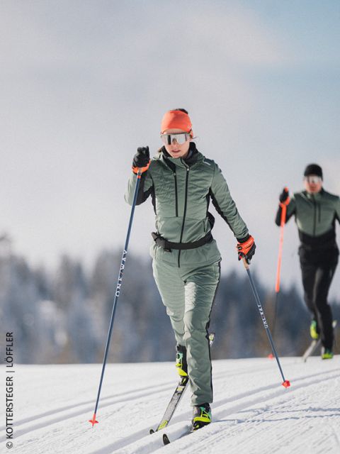 Due sciatori di fondo su una pista innevata, con attrezzatura da sport invernali e occhiali da sole, con alberi e cielo azzurro sullo sfondo.