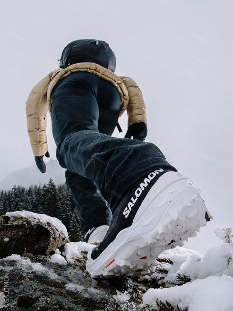 Una persona in abbigliamento invernale e scarpe Salomon cammina in salita sulla neve, con l'attenzione sulla suola della scarpa e uno sfondo montuoso innevato.