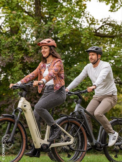 Una donna e un uomo guidano insieme una bicicletta elettrica in un parco verde, entrambi indossano caschi e abbigliamento casual, sorridono e si godono la natura.