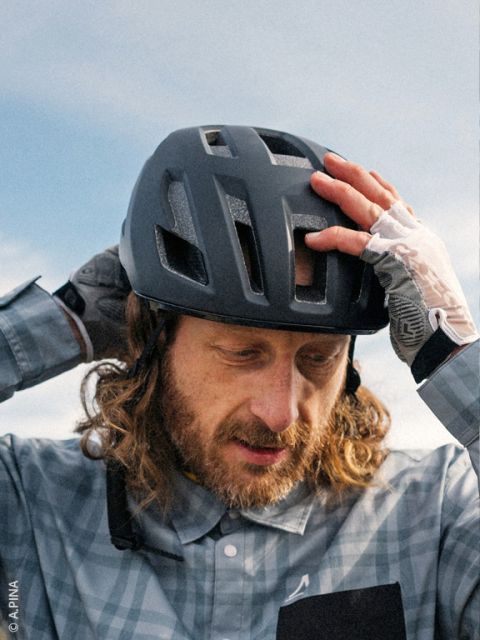 Un uomo con i capelli lunghi e la barba si sistema il casco da bici nero, indossa una camicia a quadri e guanti senza dita, sullo sfondo si vede il cielo.