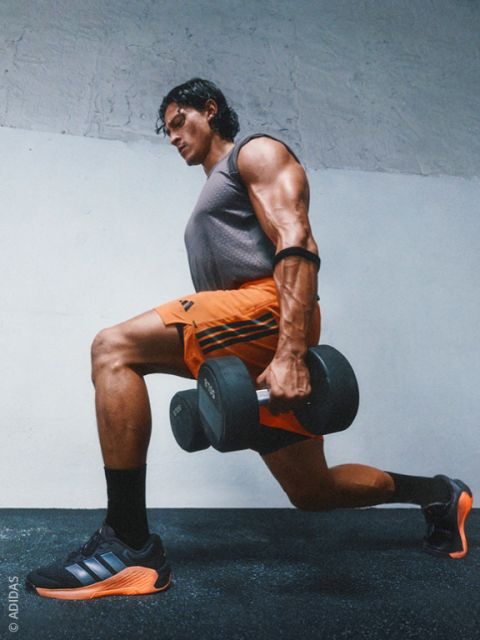 Un uomo muscoloso in abbigliamento sportivo esegue un affondo con manubri in una palestra e indossa scarpe da ginnastica Adidas nere e arancioni.