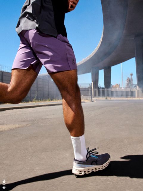 Persona che cammina su una strada soleggiata sotto un cavalcavia autostradale, con pantaloncini viola, calzini bianchi e scarpe da corsa grigie.