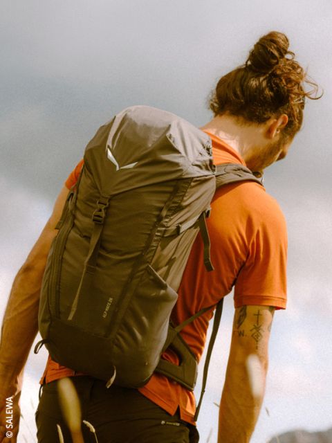 Una persona con uno chignon, una camicia arancione e uno zaino è in piedi all'aperto sotto un cielo nuvoloso e guarda in lontananza.