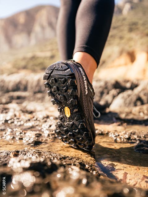 Primo piano di una persona con scarpe da trail running nere che corre su un terreno roccioso e bagnato all'aperto, con montagne sullo sfondo sfocato.