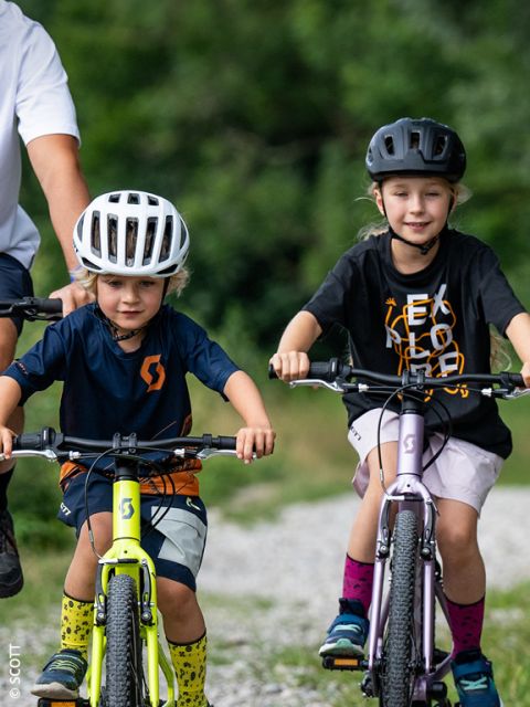 Due bambini piccoli con i caschi pedalano all'aperto su un sentiero di ghiaia, mentre un adulto è in piedi nelle vicinanze. Nello sfondo sfocato si vede del verde.