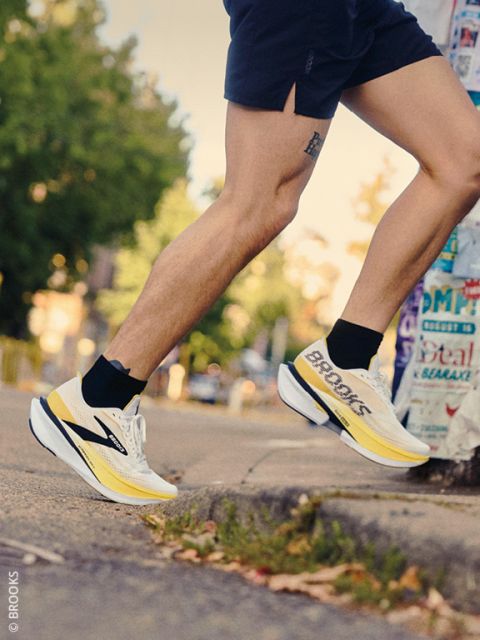 Un corridore in pantaloncini e scarpe da corsa Brooks giallo-bianche sale su un marciapiede. Sullo sfondo si vedono alberi e un palo coperto di manifesti.