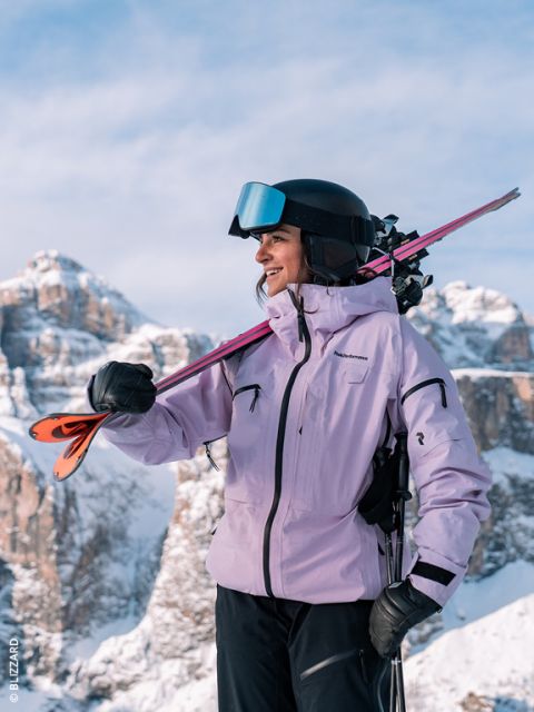 Una sciatrice con una giacca color lavanda e un casco sorride mentre porta gli s