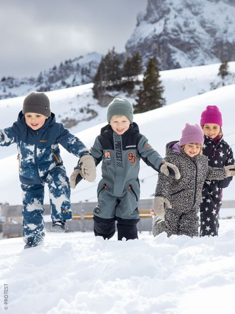Quattro bambini in abiti invernali giocano e sorridono nella neve, con montagne 