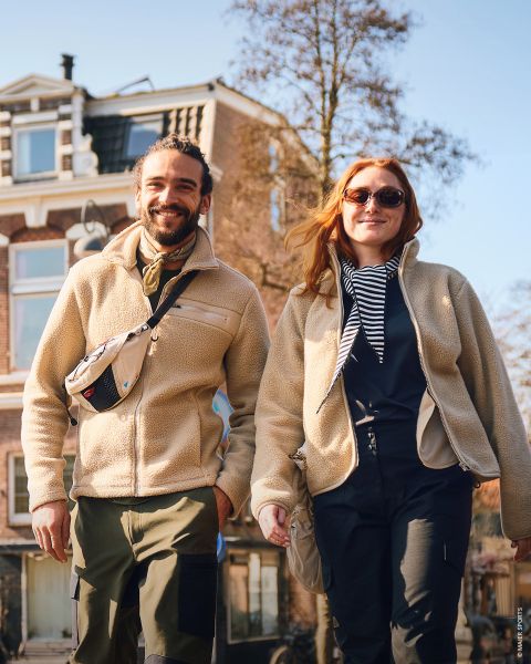 Un uomo e una donna sorridenti camminano all'aperto in una giornata di sole, vestiti con giacche e stivali casual, tenendosi per mano, con un grande edificio in mattoni sullo sfondo.