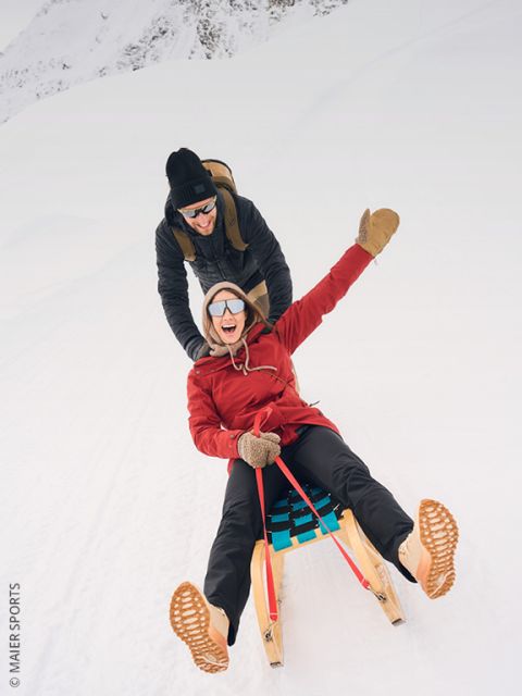 Due persone in abbigliamento invernale trainano una slitta di legno su una collina innevata, con alberi e una cima di montagna illuminata dal sole sullo sfondo. In lontananza si possono vedere altre persone.